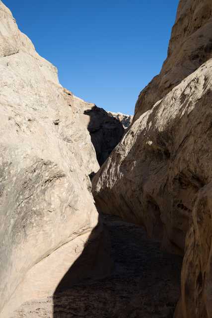 A narrow slot canyon with high, light-colored rock walls and a sandy floor.