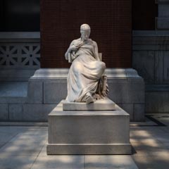 A marble statue of a seated woman in classical drapery rests on a stone pedestal in a paved courtyard, flanked by brick and stone architectural elements.