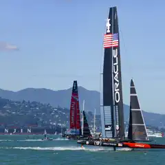 A sailboat with "ORACLE" on the sail is racing in a bay with mountains in the background.