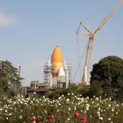 A space shuttle exhibit is displayed outdoors, surrounded by scaffolding, a crane, and a flower garden in the foreground.