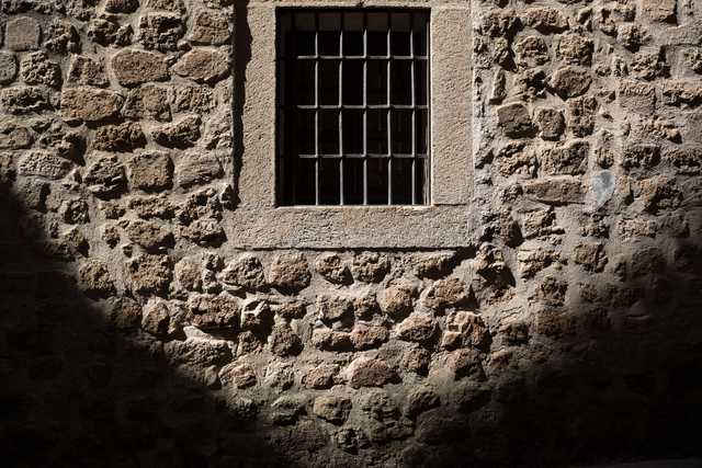 A barred window on a stone wall, with sunlight casting shadows and highlighting textures.