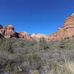 A rugged landscape features layered red rock formations under a clear blue sky, with dense greenery in the foreground.