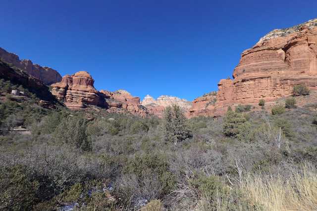 A rugged landscape features layered red rock formations under a clear blue sky, with dense greenery in the foreground.