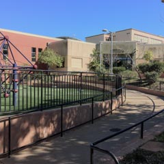 A curved sidewalk with handrails leads past a playground with a climbing structure and artificial turf, adjacent to a school building.