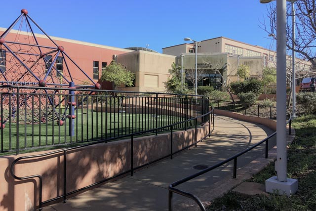A curved sidewalk with handrails leads past a playground with a climbing structure and artificial turf, adjacent to a school building.