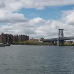 A suspension bridge spans a body of water flanked by an industrial waterfront and a row of multi-story residential buildings.
