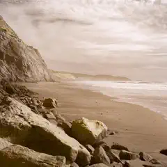 A rocky shoreline with large boulders in the foreground, a sandy beach extending toward the ocean, and a steep cliff rising on the left.