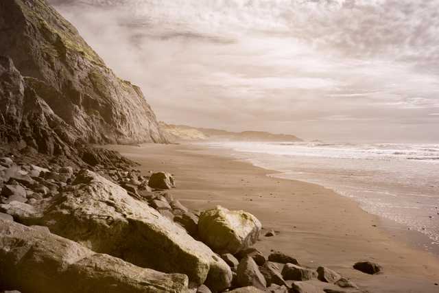 A rocky shoreline with large boulders in the foreground, a sandy beach extending toward the ocean, and a steep cliff rising on the left.