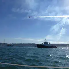 A dark military jet flies over a body of water, leaving a white smoke trail, while a tugboat and several sailboats are visible below.