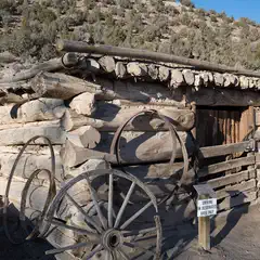 A rustic log cabin with a flat roof of stacked logs, surrounded by wooden wagon wheels, a fence, and dry, scrub-covered hills.