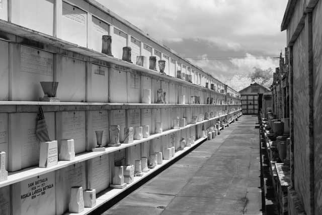 A long aisle between rows of marble crypts in a cemetery, with niches holding urns and inscribed plaques.