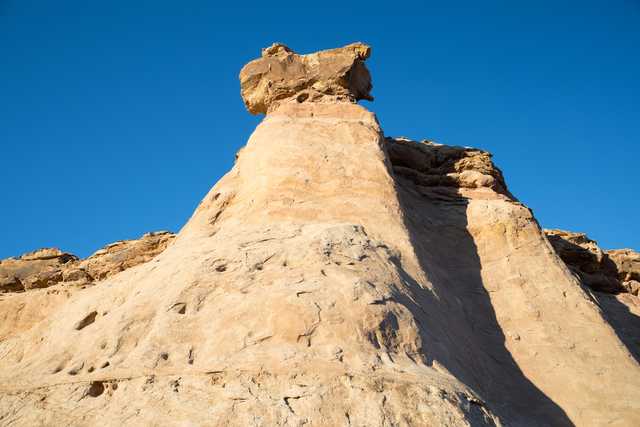 A tall, conical rock formation with a large, balanced boulder on top, set against a clear blue sky.