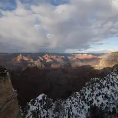A vast canyon with layered rock formations stretches into the distance, its slopes dusted with snow under a partly cloudy sky.