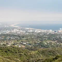 A densely vegetated hillside transitions into a suburban area with scattered residential buildings, leading to a coastal cityscape and a distant shoreline.