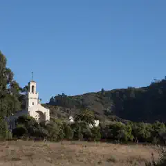 A white church with a bell tower stands among trees on a hillside under a clear blue sky.