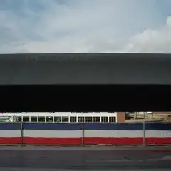 A large aircraft with AIRFORCE lettering on its fuselage is positioned above a building with a red, white, and blue striped fence.