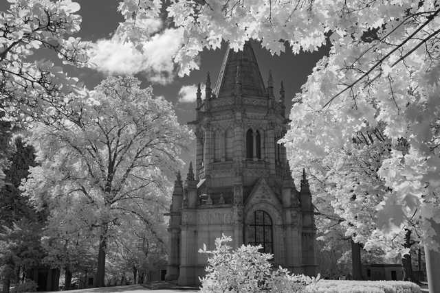 A stone mausoleum with ornate Gothic detailing is framed by leafy trees with bright foliage, set in a cemetery landscape.
