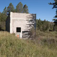 A concrete block building with barred windows and a metal door stands among tall grass and evergreen trees.