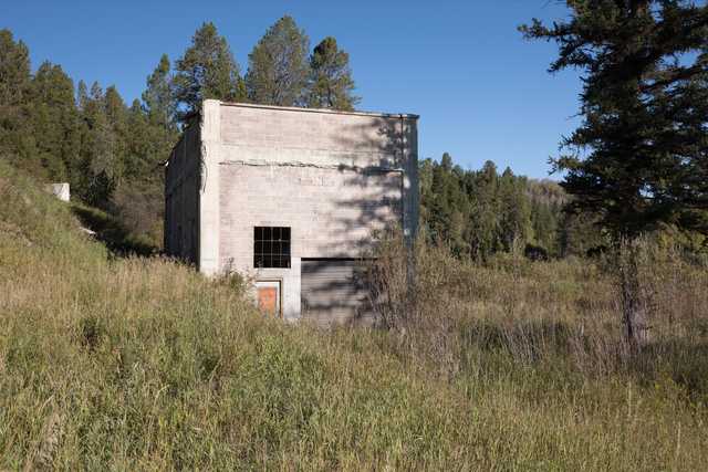 A concrete block building with barred windows and a metal door stands among tall grass and evergreen trees.