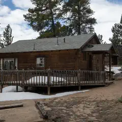 A wooden cabin with a front porch stands among pine trees, with snow patches on the ground.