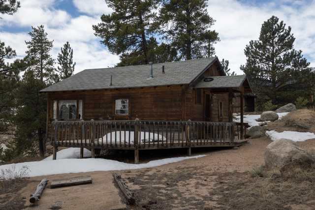 A wooden cabin with a front porch stands among pine trees, with snow patches on the ground.
