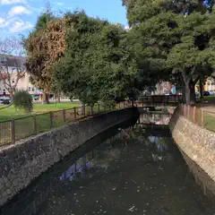A bridge over a small creek. The creek is narrow and flowing between stone walls. Trees line the banks of the creek. Buildings can be seen in the background.