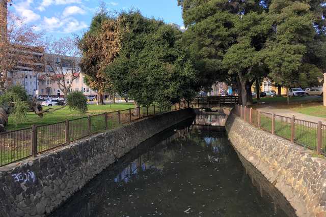 A bridge over a small creek. The creek is narrow and flowing between stone walls. Trees line the banks of the creek. Buildings can be seen in the background.