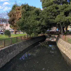 A bridge over a small creek. The creek is narrow and flowing between stone walls. Trees line the banks of the creek. Buildings can be seen in the background.