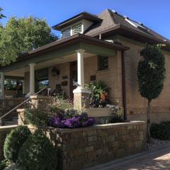 A small house made of light brown bricks, featuring a covered porch with white columns and surrounded by well-manicured landscaping.