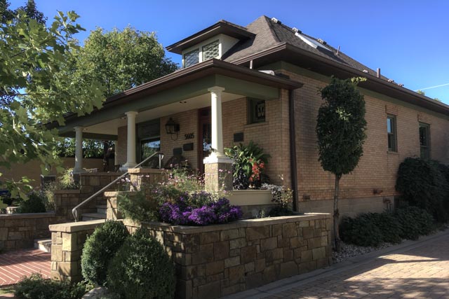 A small house made of light brown bricks, featuring a covered porch with white columns and surrounded by well-manicured landscaping.