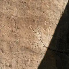 An ancient rock surface features various petroglyphs, including spirals, zigzags, and animal figures.
