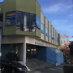 A school building featuring a rainbow flag in its window is situated on a street lined with parked cars, trees, and power lines.