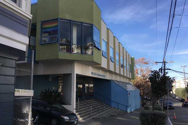 A school building featuring a rainbow flag in its window is situated on a street lined with parked cars, trees, and power lines.