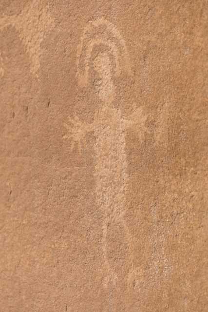 A petroglyph on a reddish-brown rock face depicts a stylized human figure with an elongated body.